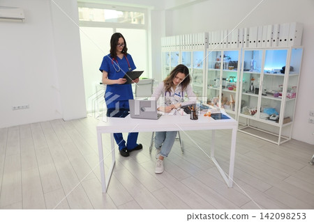 Two female medical professionals working together in bright clinic office, one standing with clipboard while other writes notes beside laptop, cabinets, files, and lab supplies Two female medical professionals working together in bright clinic office, one standing with clipboard while other writes notes beside laptop, cabinets, files, and lab supplies 142098253