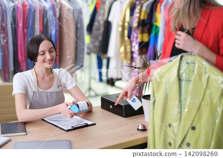 Smiling boutique clerk processing contactless payment for customer choosing colorful garments beside clothing rack and checkout desk in bright fashion store interior Smiling boutique clerk processing contactless payment for customer choosing colorful garments beside clothing rack and checkout desk in bright fashion store interior 142098260