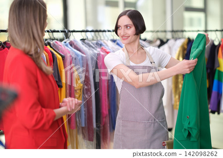 Fashion store assistant presenting green dress to shopper beside colorful garment rack inside bright boutique with friendly personal service and stylish retail atmosphere 142098262