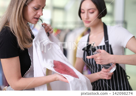 Two women in aprons inspect stained white fabric while discussing cleaning treatment, with one holding spray bottle and other examining red spill mark in bright indoor workspace Two women in aprons inspect stained white fabric while discussing cleaning treatment, with one holding spray bottle and other examining red spill mark in bright indoor workspace 142098274