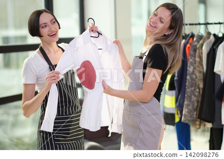 Two smiling women in aprons holding white shirt with red heart stain during laundry or garment care task beside clothing rack in bright studio with casual teamwork and creative repair concept Two smiling women in aprons holding white shirt with red heart stain during laundry or garment care task beside clothing rack in bright studio with casual teamwork and creative repair concept 142098276