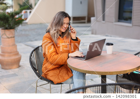 Woman working on laptop and talking on phone, in outdoor cafe, with laptop, phone headset, coffee cup, table, from a side view perspective, with orange, black accents. Woman working on laptop and talking on phone, in outdoor cafe, with laptop, phone headset, coffee cup, table, from a side view perspective, with orange, black accents. 142098301