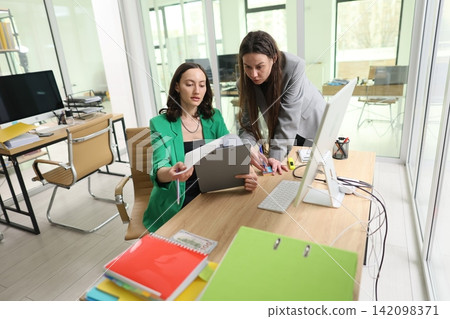Two businesswomen reviewing documents together in bright modern office, one seated holding clipboard while colleague leans over desk to discuss work, teamwork and focused planning Two businesswomen reviewing documents together in bright modern office, one seated holding clipboard while colleague leans over desk to discuss work, teamwork and focused planning 142098371