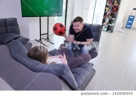 Smiling man holding red soccer ball while sitting on modern chair and facing relaxed person on sofa in bright living room with television, shelves, pale floor, and casual indoor play atmosphere Smiling man holding red soccer ball while sitting on modern chair and facing relaxed person on sofa in bright living room with television, shelves, pale floor, and casual indoor play atmosphere 142098393