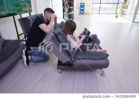 Father and young daughter spending quiet time on modern sofa while girl uses tablet and man kneels beside her in bright living room, showing concern, supervision and family attention at home Father and young daughter spending quiet time on modern sofa while girl uses tablet and man kneels beside her in bright living room, showing concern, supervision and family attention at home 142098400
