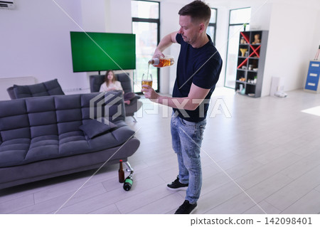 Man pours amber beverage into wine glass inside bright modern living room, with blurred woman resting on sofa, bottles on floor, soft daylight and relaxed casual home gathering Man pours amber beverage into wine glass inside bright modern living room, with blurred woman resting on sofa, bottles on floor, soft daylight and relaxed casual home gathering 142098401