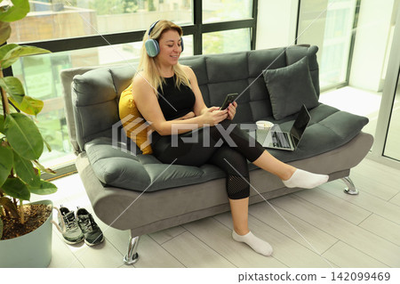Relaxed woman wearing headphones using smartphone while sitting on modern sofa with laptop nearby in bright home interior, enjoying calm music, casual leisure, and quiet indoor comfort Relaxed woman wearing headphones using smartphone while sitting on modern sofa with laptop nearby in bright home interior, enjoying calm music, casual leisure, and quiet indoor comfort 142099469