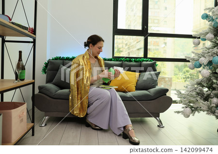 Woman in gold sequined shawl sorting cards on sofa beside decorated Christmas tree in bright modern living room with city window light, festive home setting, gift preparation, calm seasonal mood Woman in gold sequined shawl sorting cards on sofa beside decorated Christmas tree in bright modern living room with city window light, festive home setting, gift preparation, calm seasonal mood 142099474