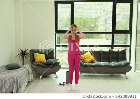 Woman in bright pink activewear doing home stretching exercise on yoga mat inside modern living room with large windows, soft daylight, sofa, pillows, dumbbells, calm indoor fitness routine Woman in bright pink activewear doing home stretching exercise on yoga mat inside modern living room with large windows, soft daylight, sofa, pillows, dumbbells, calm indoor fitness routine 142099911