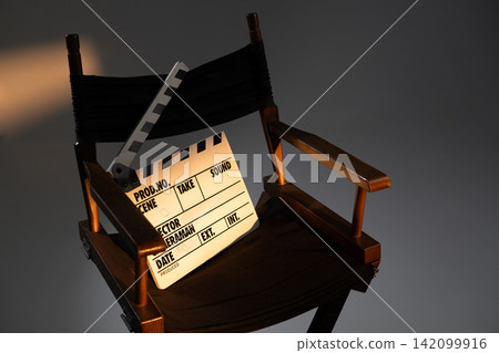 Close up of a director chair with a movie clapperboard lit by warm studio light in a dark background Close up of a director chair with a movie clapperboard lit by warm studio light in a dark background 142099916