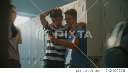 Two Cheerful Teenage Classmates Stand by School Lockers, Sharing Smartphone and Laughing Together Two Cheerful Teenage Classmates Stand by School Lockers, Sharing Smartphone and Laughing Together 142100220