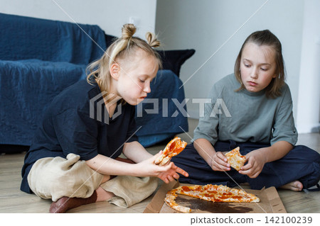 Cheerful young children sitting on floor in living room and eating pizza at home Cheerful young children sitting on floor in living room and eating pizza at home 142100239
