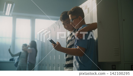 Friendly Teenage Classmates Relax by Lockers while One Shares Something Entertaining on Smartphone 142100295