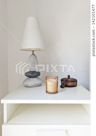 Stone table lamp, glowing ribbed candle holder and dark glass decorative container on white cabinet against neutral wall 142101477
