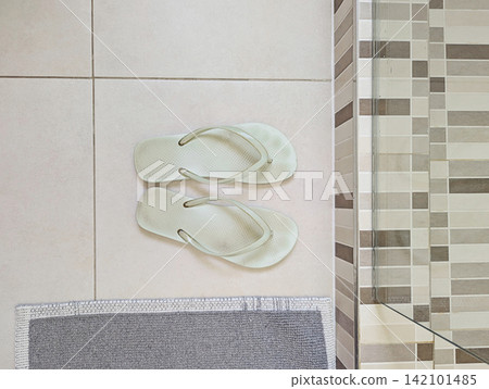 Mint flip flops on neutral tiled bathroom floor beside glass shower enclosure and gray bath mat, clean simple home interior Mint flip flops on neutral tiled bathroom floor beside glass shower enclosure and gray bath mat, clean simple home interior 142101485