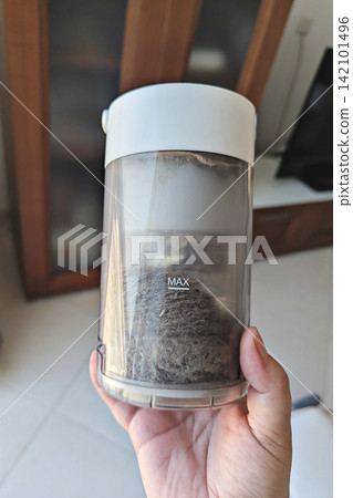 Transparent vacuum dust bin filled with hair, lint and dirt, held by hand indoors before emptying and cleaning appliance Transparent vacuum dust bin filled with hair, lint and dirt, held by hand indoors before emptying and cleaning appliance 142101496