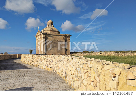 Kappella San Nikolaw chapel beside paved path and traditional Maltese stone walls, green fields and bright blue sky with scattered clouds in rural Malta 142101497