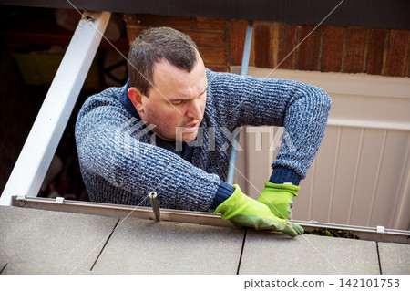 Man on a ladder cleaning fallen autumn leaves and moss from house roof gutter. Home maintenance, gutter cleaning Man on a ladder cleaning fallen autumn leaves and moss from house roof gutter. Home maintenance, gutter cleaning 142101753