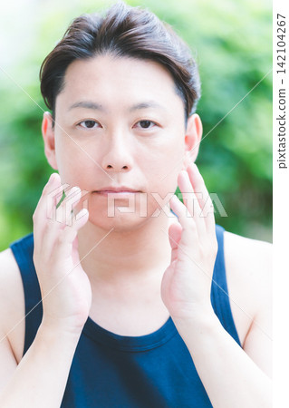 A man in his 40s performing botanical skincare outdoors, applying facial care with both hands to his cheeks. 142104267
