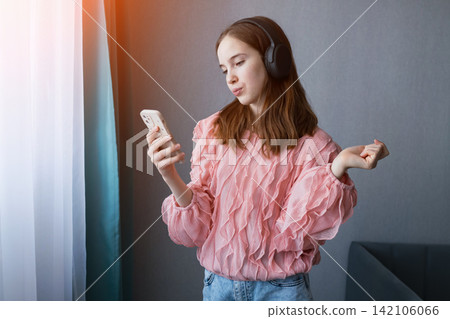 Young female enjoying her leisure time at home, wearing headphones and actively engaging with her mobile phone, embodying youth culture and digital connection 142106066