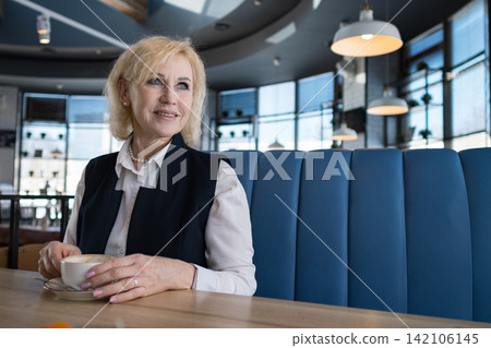 Senior Businesswoman Enjoying Quiet Cafe Moment Seated In Blue Booth With Natural Light, Smiling Softly While Holding Cup And Resting Hands On Table, Casual Corporate Attire With Blazer And Blouse, 142106145
