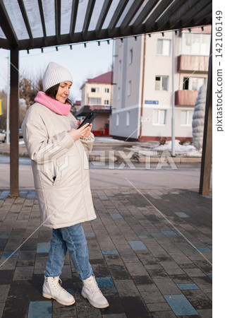 Pregnant woman in winter coat and hat outdoors holding a phone near apartment buildings Pregnant woman in winter coat and hat outdoors holding a phone near apartment buildings 142106149