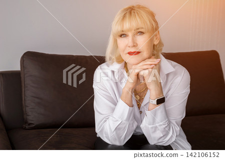 Senior woman with blonde hair and red lipstick sitting on a dark couch, expressing confidence and elegance in a portrait promoting mature beauty, wisdom, and retirement 142106152