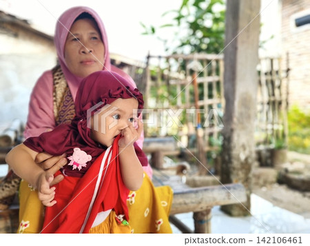 Indonesian grandmother and granddaughter wearing hijab sitting outside their village home. 142106461