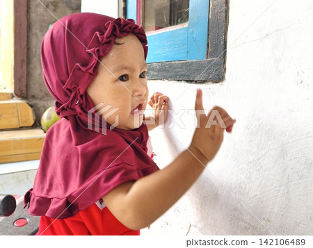 Cute Muslim baby in burgundy hijab looking curious with blurred mother Cute Muslim baby in burgundy hijab looking curious with blurred mother 142106489