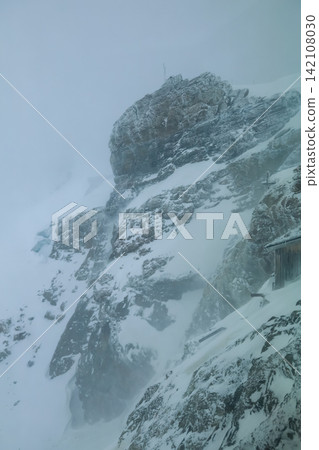 Snow Covered Mountain Cliff with Alpine Hut Shrouded in Fog and Mist Snow Covered Mountain Cliff with Alpine Hut Shrouded in Fog and Mist 142108030