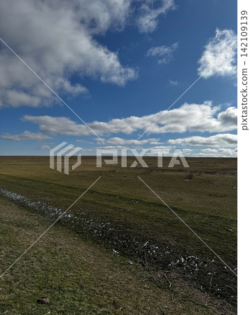 Wide grassy plain under blue sky with scattered white clouds and sunlight. Wide grassy plain under blue sky with scattered white clouds and sunlight. 142109139