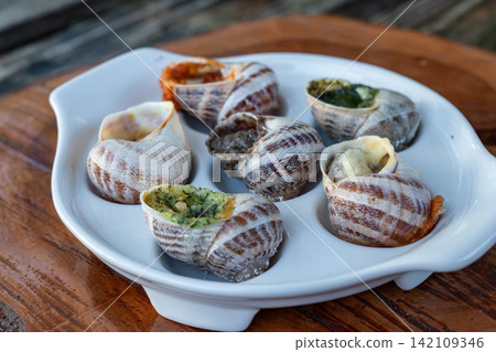Baked stuffed mollusks are presented with green herb pesto and red spicy tomato sauces inside special white tray on timber background near metal fork outdoors. Baked stuffed mollusks are presented with green herb pesto and red spicy tomato sauces inside special white tray on timber background near metal fork outdoors. 142109346