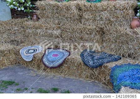 Decorative village location features multiple dry agriculture bundles arranged as seating bench with colorful round textile items outdoors under day light. Decorative village location features multiple dry agriculture bundles arranged as seating bench with colorful round textile items outdoors under day light. 142109365