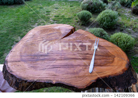 Two-tined metallic dining utensil rests on varnished natural timber platform section outdoors near green bushes under daylight during summer culinary exhibition. 142109366