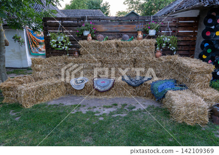 Decorative village location features multiple dry agriculture bundles arranged as sofa with colorful handmade rugs standing near brown timber barrier in green garden. Decorative village location features multiple dry agriculture bundles arranged as sofa with colorful handmade rugs standing near brown timber barrier in green garden. 142109369