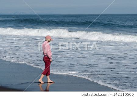 Woman walks barefoot along Pacific shoreline on black volcanic sand at Khalaktyrsky Beach, Kamchatka, Russia. Travel, nature tourism, active lifestyle, coastal recreation, freedom, summer vacation. 142110500