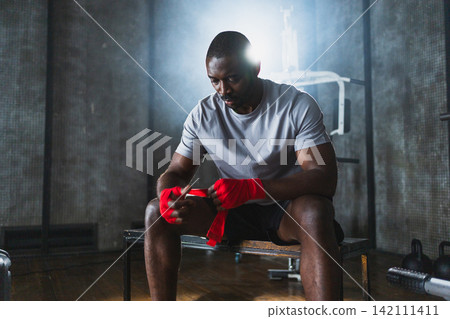 Fitness workout in gym. African American man fighter preparing for fight wrapping hands with red boxing wraps sports protective bandages in gym. Strong man ready for fight boxing sparring training Fitness workout in gym. African American man fighter preparing for fight wrapping hands with red boxing wraps sports protective bandages in gym. Strong man ready for fight boxing sparring training 142111411