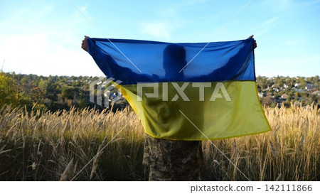 Military man in uniform stands with raised over head flag of Ukraine at countryside. Male ukrainian army soldier with lifted national banner in honor of victory against russian aggression. End of war Military man in uniform stands with raised over head flag of Ukraine at countryside. Male ukrainian army soldier with lifted national banner in honor of victory against russian aggression. End of war 142111866
