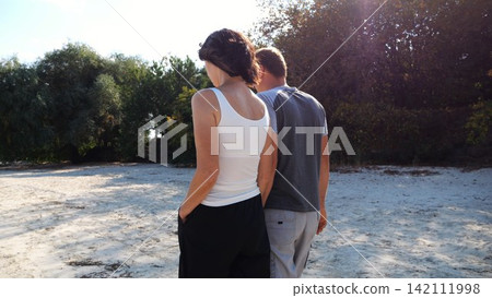 Happy pair going along coast at sunny day. Romantic couple holding hands of each other and walking on sandy beach. Lovers spending time together at resort. Concept of summer vacation or holiday 142111998