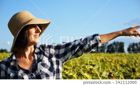 Happy smiling female farmer looks into sunflower field and enjoys harvest. Portrait of adult beautiful agronomist in straw hat with yellow plantation at background. Concept of agricultural business Happy smiling female farmer looks into sunflower field and enjoys harvest. Portrait of adult beautiful agronomist in straw hat with yellow plantation at background. Concept of agricultural business 142112000