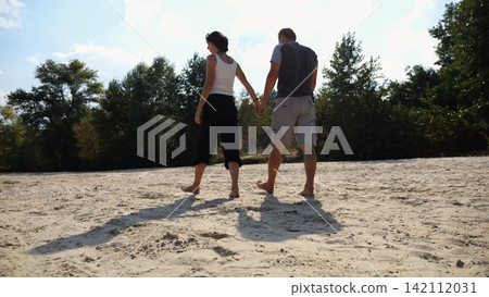 Romantic couple holding hands of each other and walking at coastline. Happy pair going along seashore at sunny day. Lovers spending time together at resort. Concept of vacation or holiday. Slow motion Romantic couple holding hands of each other and walking at coastline. Happy pair going along seashore at sunny day. Lovers spending time together at resort. Concept of vacation or holiday. Slow motion 142112031