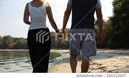 Romantic couple holding hands of each other and walking at coastline. Happy pair going along seashore at sunny day. Lovers spending time together at resort. Concept of vacation or holiday. Slow motion Romantic couple holding hands of each other and walking at coastline. Happy pair going along seashore at sunny day. Lovers spending time together at resort. Concept of vacation or holiday. Slow motion 142112037