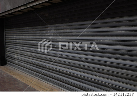 A store with its shutters down. It gives the impression of being closed or temporarily shut down. The shutters are made of metal. A store with its shutters down. It gives the impression of being closed or temporarily shut down. The shutters are made of metal. 142112237
