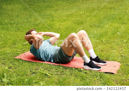 Active man doing an abs crunch exercise on a mat outdoors. Fitness workout and training on green grass in the park for a healthy lifestyle. Active man doing an abs crunch exercise on a mat outdoors. Fitness workout and training on green grass in the park for a healthy lifestyle. 142112437