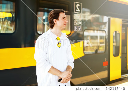 man stands on a station platform waiting with his suitcase. The passenger is ready for a train travel journey and transportation to his destination. man stands on a station platform waiting with his suitcase. The passenger is ready for a train travel journey and transportation to his destination. 142112440