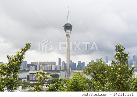 Macau Tower surrounded by green trees with a city skyline and skyscrapers under a cloudy sky. Macau Tower surrounded by green trees with a city skyline and skyscrapers under a cloudy sky. 142113151