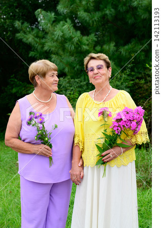 Two elderly Russian sisters aged 73 and 79 years old holding flower bouquets while smiling and standing together in a rural village garden. Two elderly Russian sisters aged 73 and 79 years old holding flower bouquets while smiling and standing together in a rural village garden. 142113193