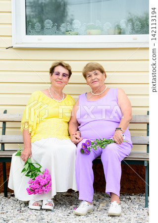 Two elderly Russian sisters aged 73 and 79 years old sitting on a wooden bench holding hands and flower bouquets next to a house in a village. 142113194