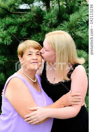 Young 17 years old Russian granddaughter kissing her elderly 73 years old grandmother on the cheek while hugging in front of a pine tree. Young 17 years old Russian granddaughter kissing her elderly 73 years old grandmother on the cheek while hugging in front of a pine tree. 142113236