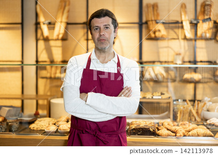 Positive adult bakery salesman in apron looking at the camera during working day in bakehouse. Cooking, culinary and bakery concept Positive adult bakery salesman in apron looking at the camera during working day in bakehouse. Cooking, culinary and bakery concept 142113978